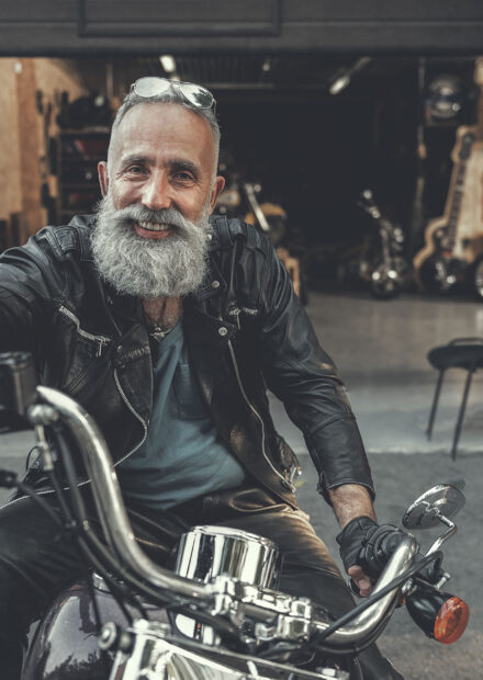 Cheerful mature bearded biker is sitting on motorcycle and holding handles. He looking at camera with bright smile. Portrait. Copy space